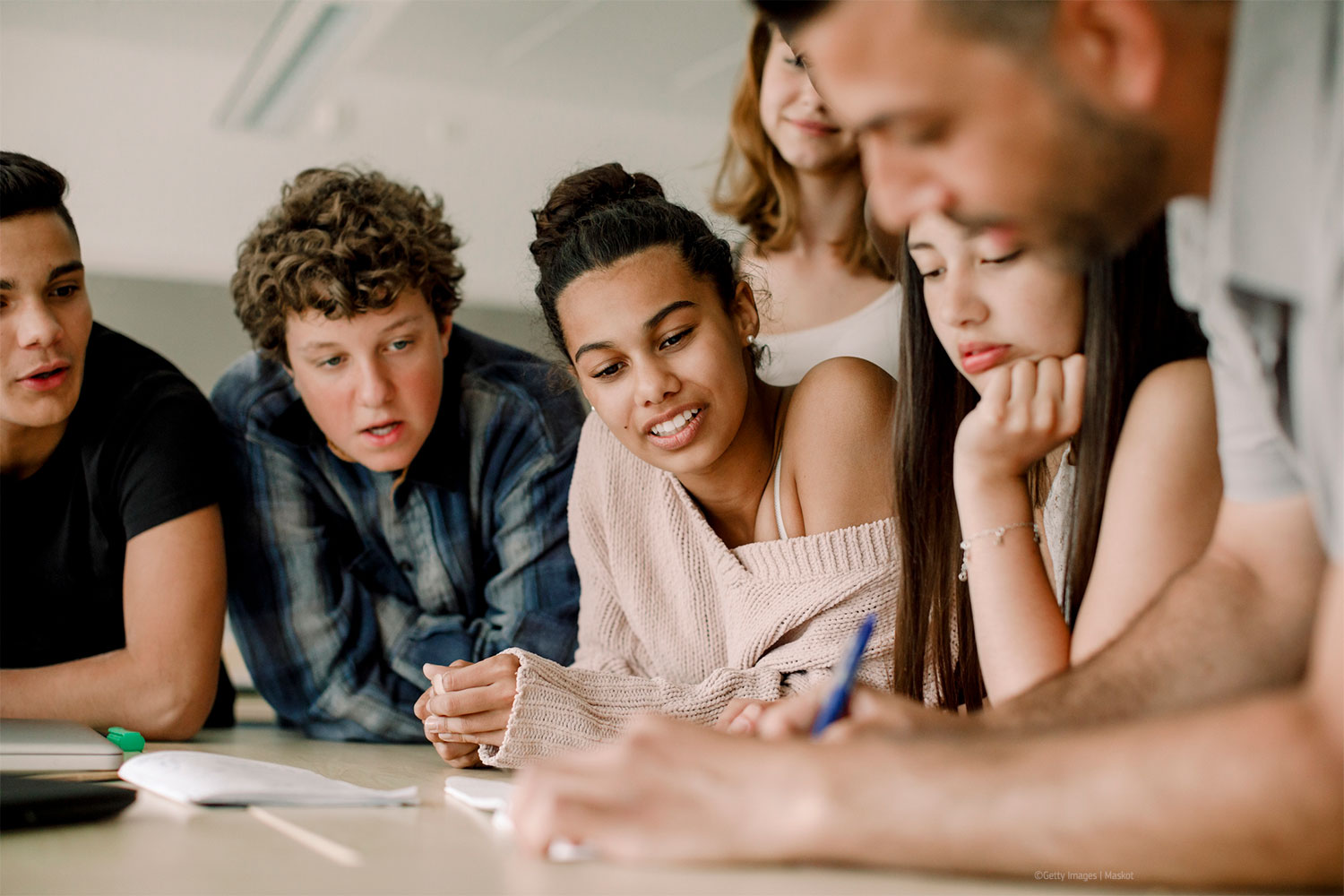 Teacher writing in book while teenage students leaning over table in classroom